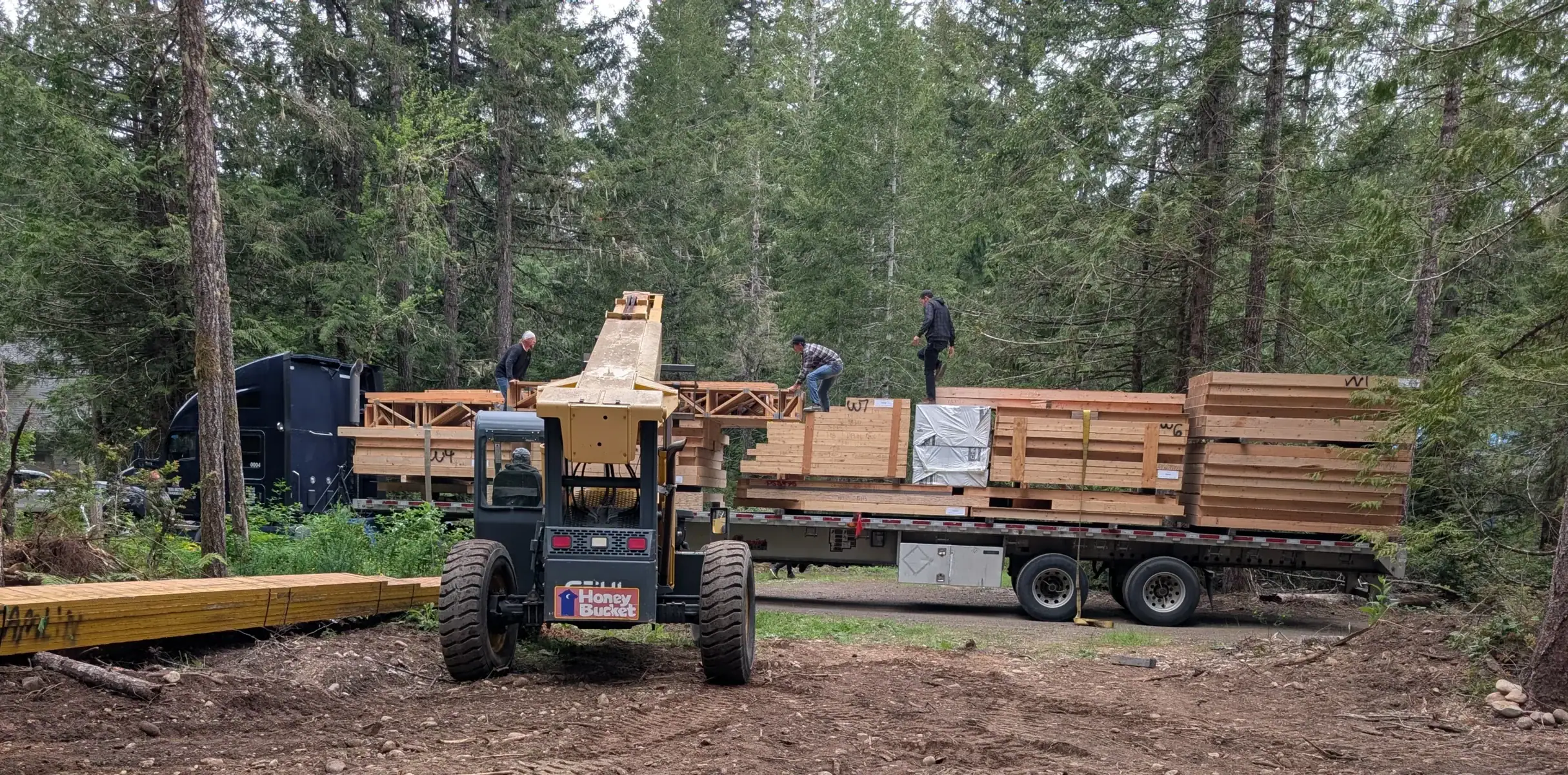 Heavy equipment unloading a prefabricated home package from a flatbed trailer on a forested Washington State building site.