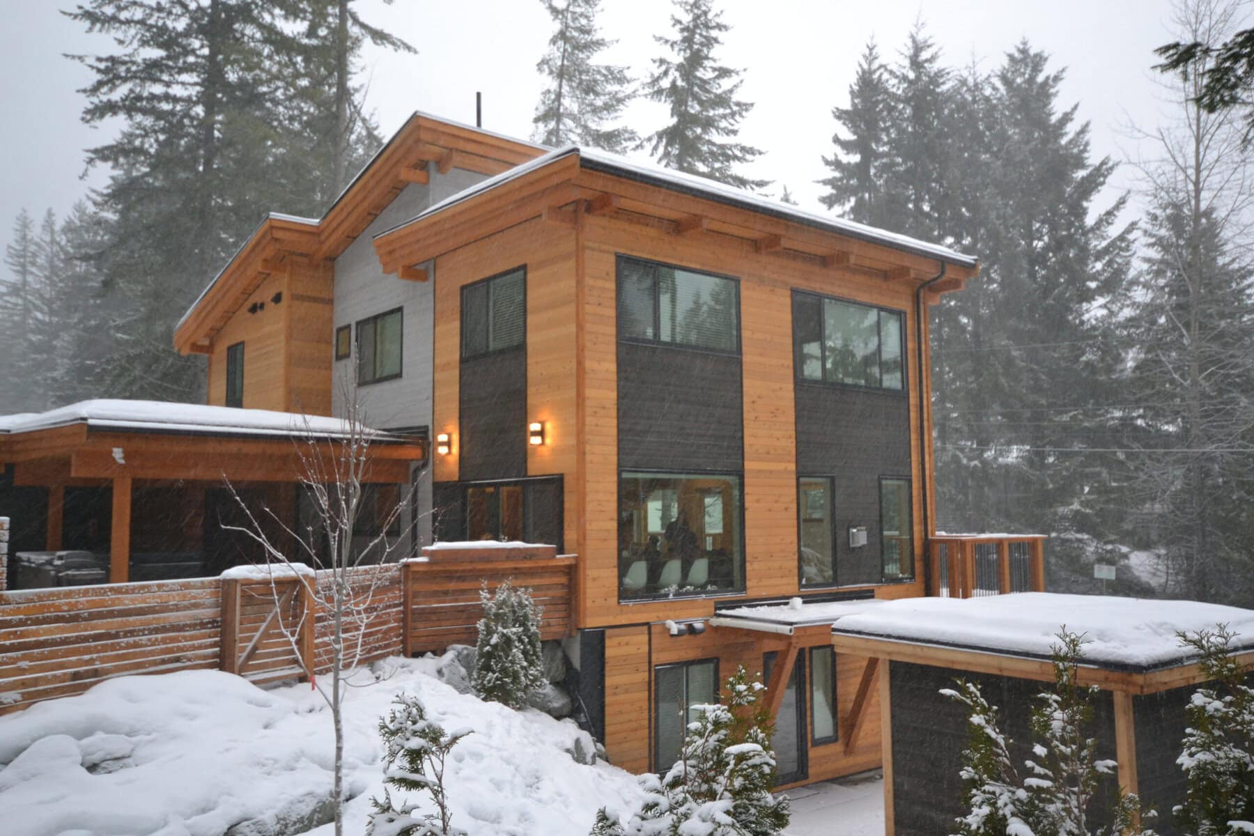 Alpine-Meadows Snow covered contemporary timber frame home in a forest, with cedar siding, dark exterior panels, large windows and layered rooflines.