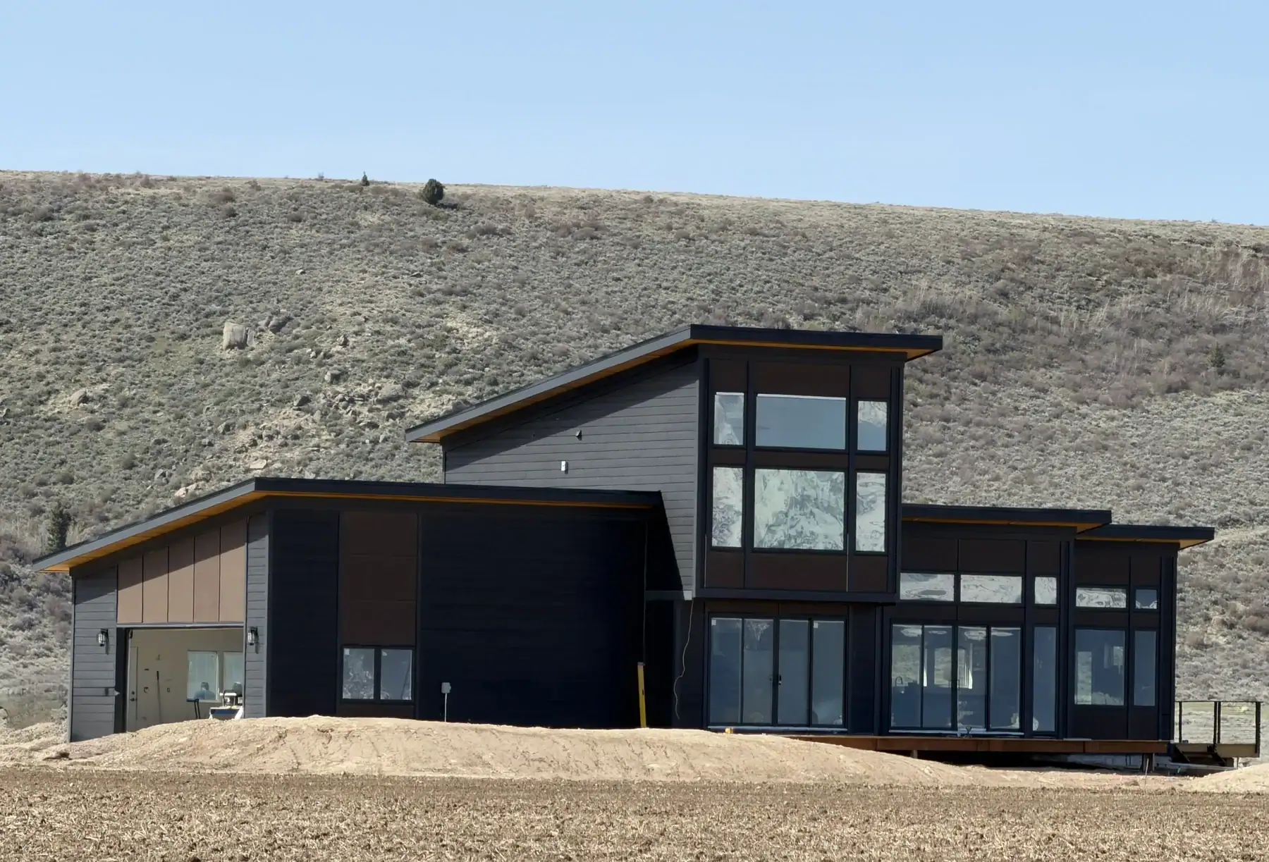 Contemporary dark sided custom home with large windows and angled rooflines, set against a dry, shrub covered hillside in Idaho.